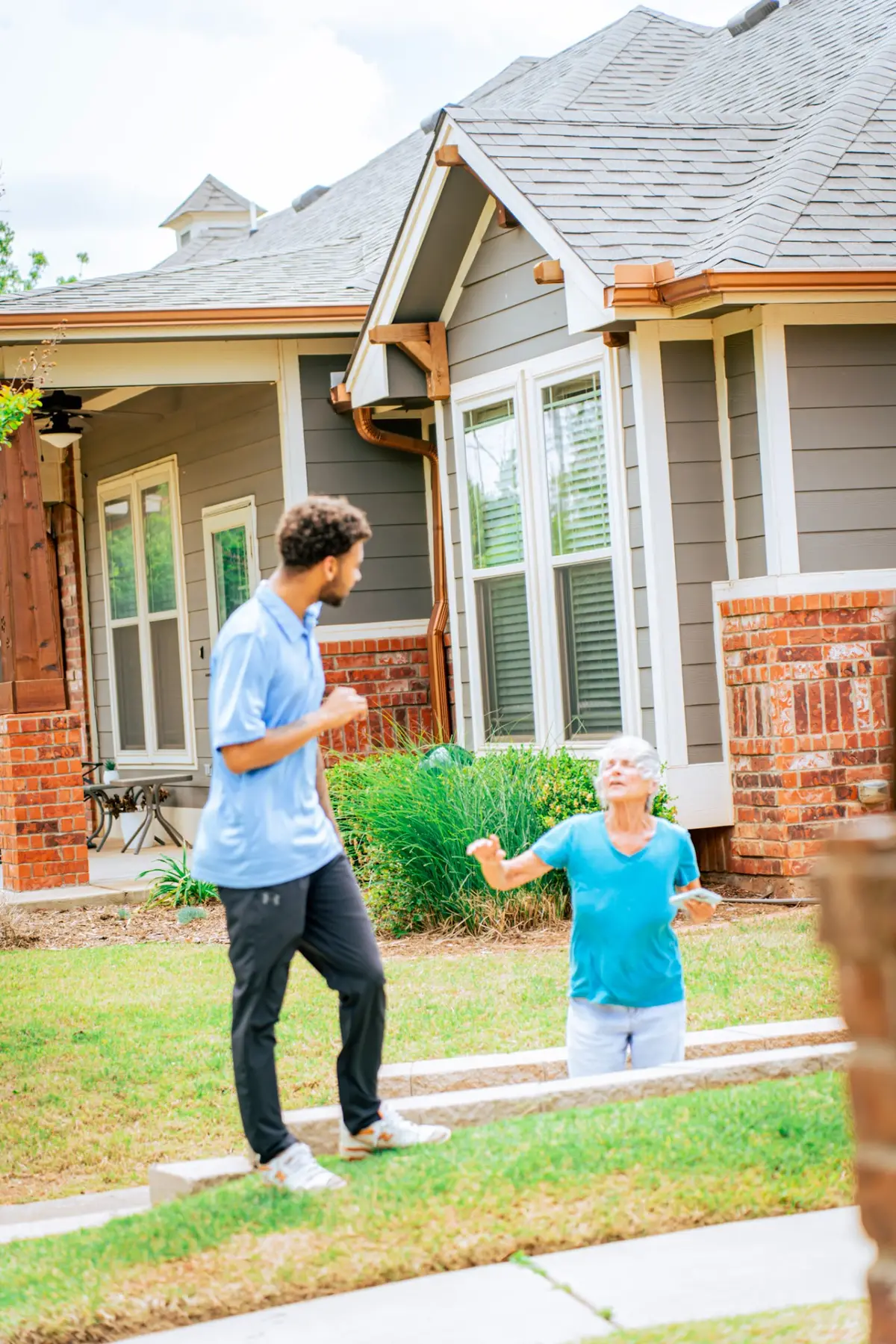 A man in a light blue polo faces an older woman gesturing beside outdoor steps. Behind them is a house with gray siding, brickwork, a shingled roof and copper-colored gutters.