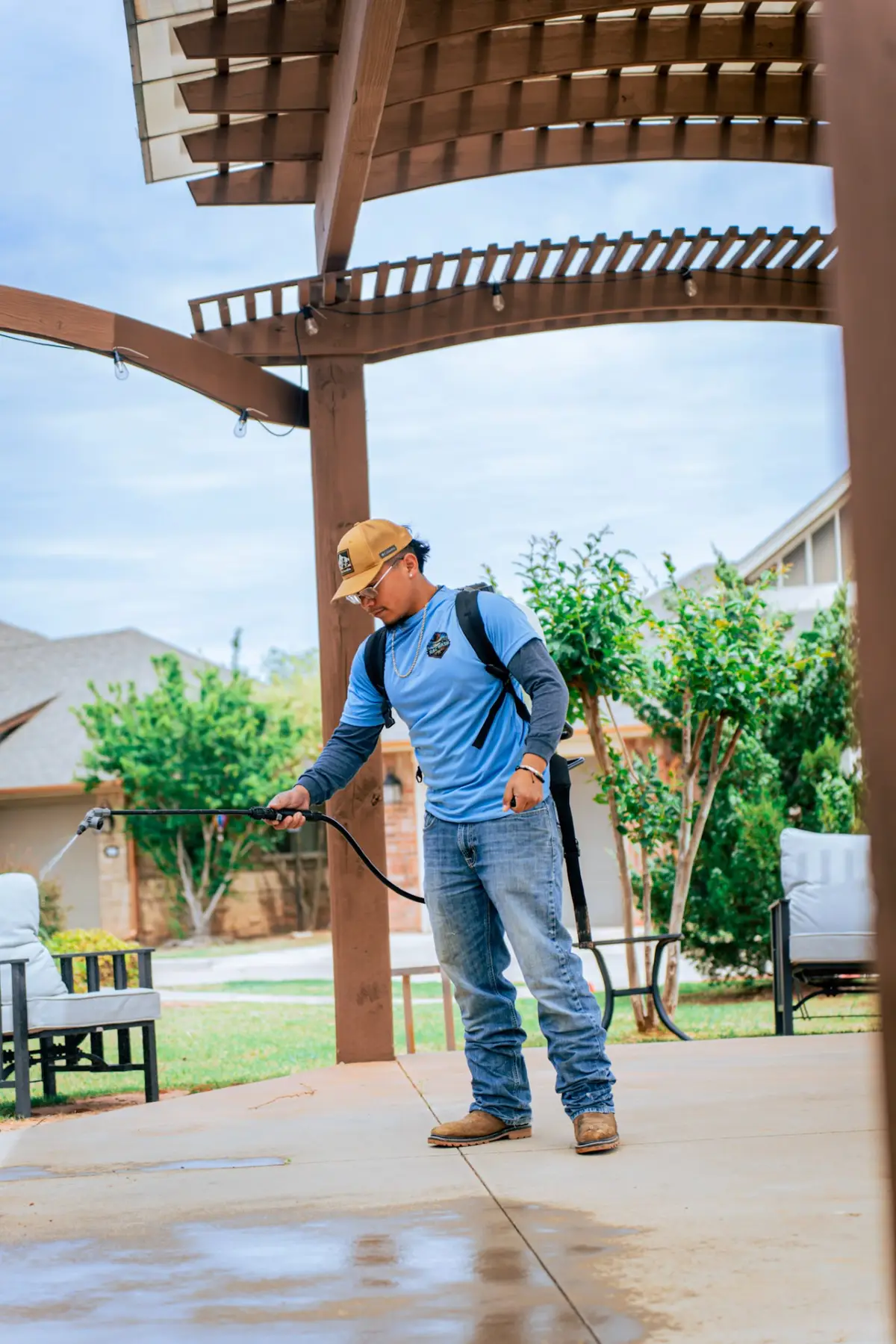 A worker wearing a backpack sprayer directs a spray wand beside patio seating beneath a wooden pergola.