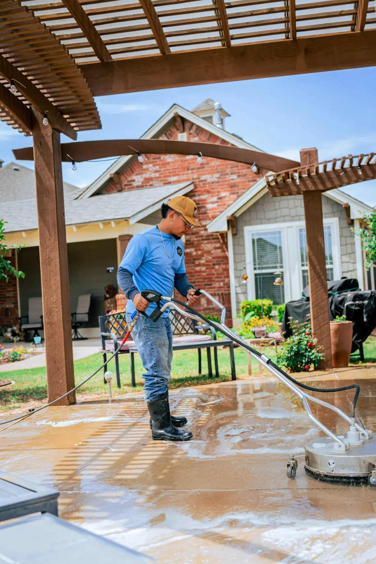 A worker in a blue shirt and rubber boots operates a wheeled surface cleaner on a wet backyard patio beneath a wooden pergola.